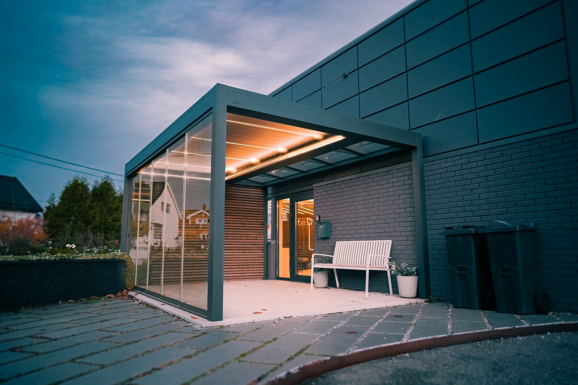 Pergola festet til siden av et hus med glassvegger, glass tak og vakker belysning på en uteplass i bakgården, bygget av uteDESIGN