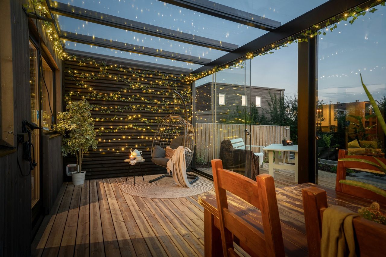 Tak pergola – 9 Bildet av en innglasset pergola med glasstak. Den er innredet med spisebord og stol i hjørnet. På den ene husveggen er det pyntet med lys.