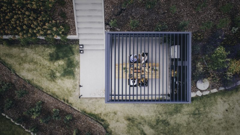 Bildet ovenfra av en pergola med lamelltak. Rundt er det en hage og inni sitter det mennesker.