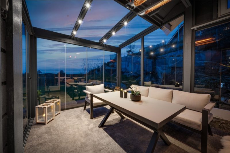 Bildet innenfra en pergola med skråtak. Det er glass på tak og vegger. Ute er det kveld og inne er det lyst med nydlige møbler.
