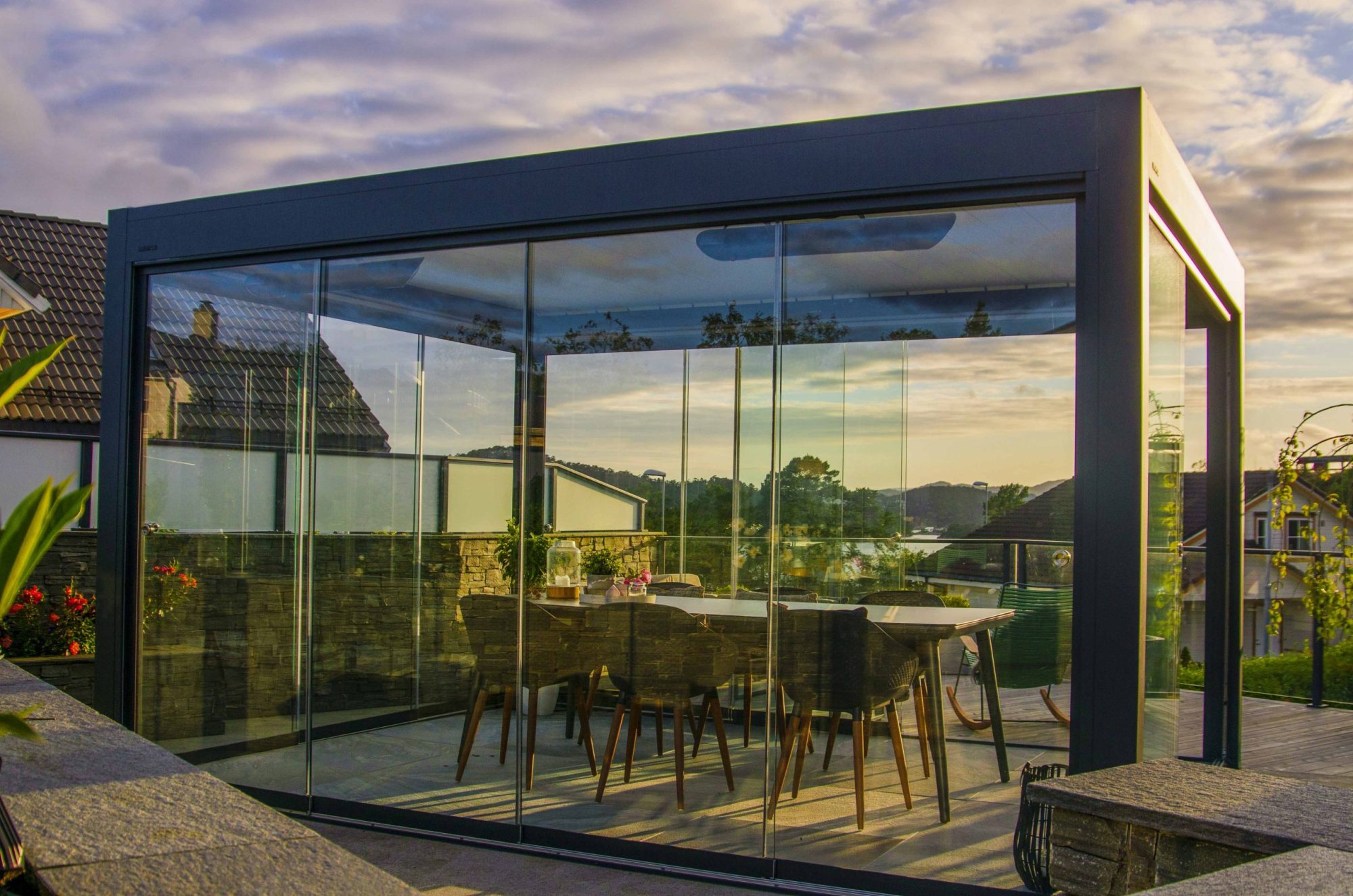 En innglasset pergola, midt på en terrasse. Det er nydelig solnedgang og pergolaen er innredet med en spiseplass.