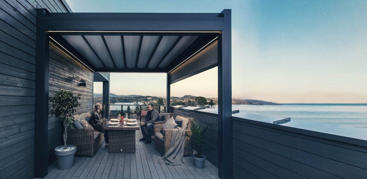 Bildet av en pergola på en takterrasse. Det er to personer som sitter inni og har en samtale, med nydelig sjøutsikt bak dem.