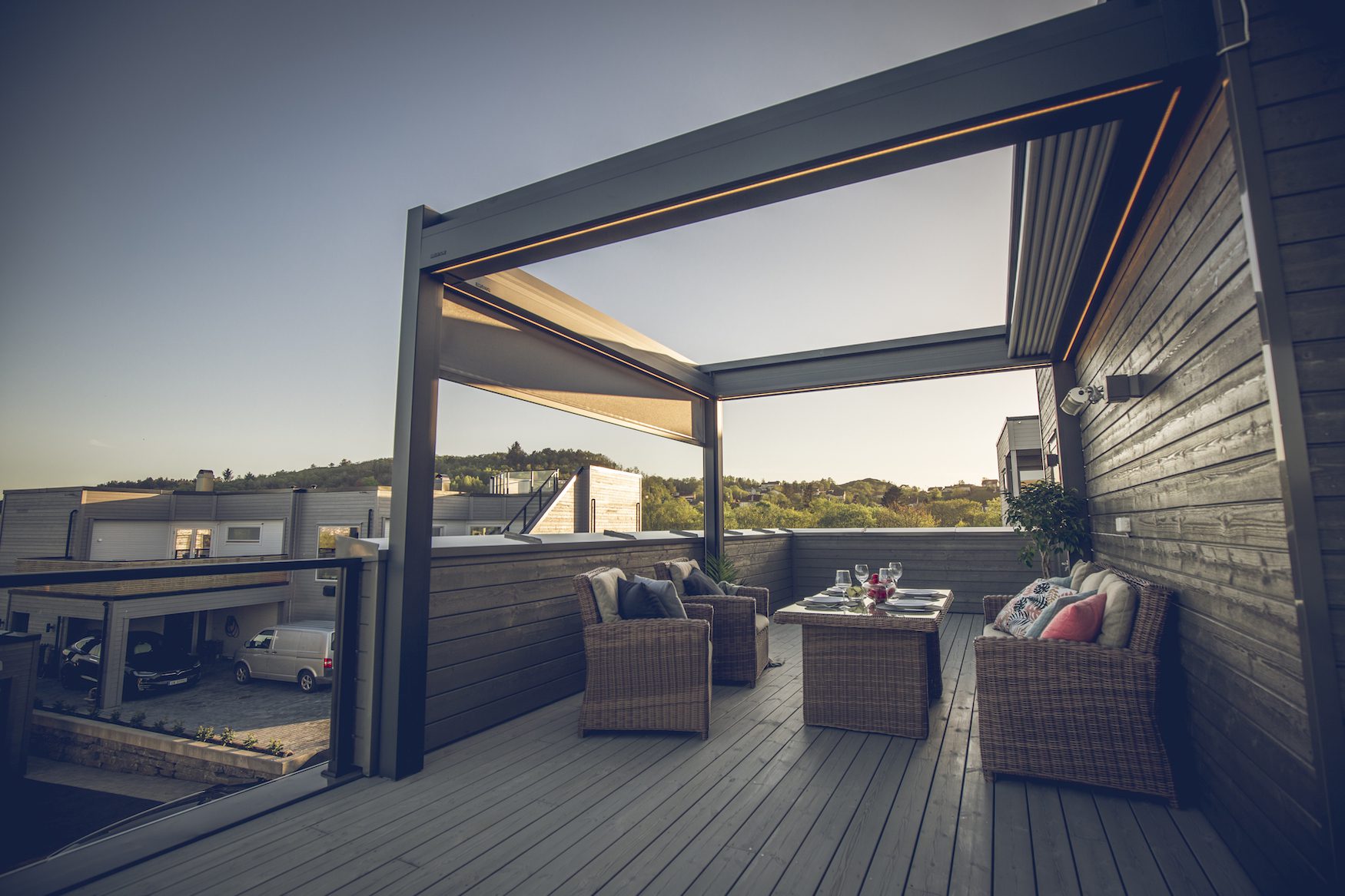 Tak pergola – 3 Bildet av en pergola i solnedgang med folddetak. Taket er tatt inn, og det er innredet med utemøbler.