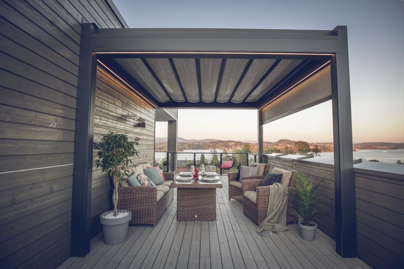 Bildet av en pergola med foldetak, hvor taket er lukket. Det er nydelig utsikt bak pergolaen og den er innredet.