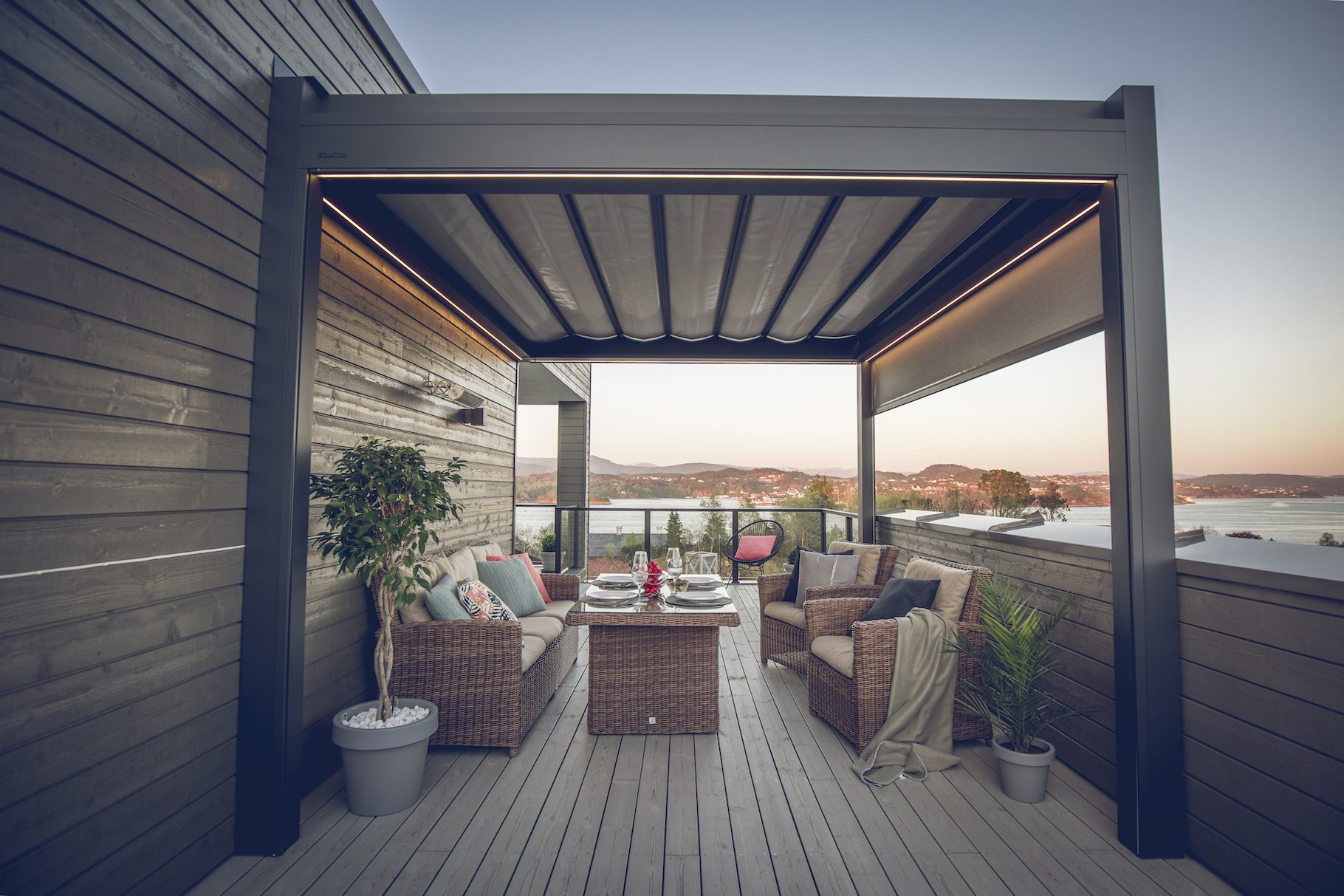 Tak pergola – 4 Bildet av en pergola med foldetak, hvor taket er lukket. Det er nydelig utsikt bak pergolaen og den er innredet.