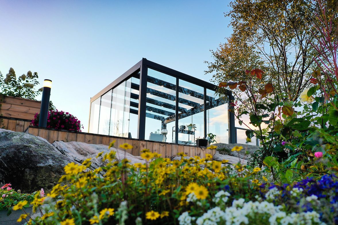 Utestue i glass på en terrasse. Bildet er tatt nedenfra og oppover, med mye blomster i front.